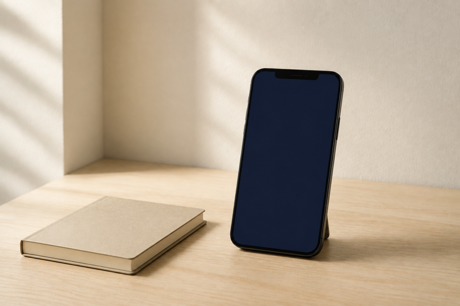 明るい木製のデスクに立てられたスマートフォンと小さなノート。窓から自然光が差している